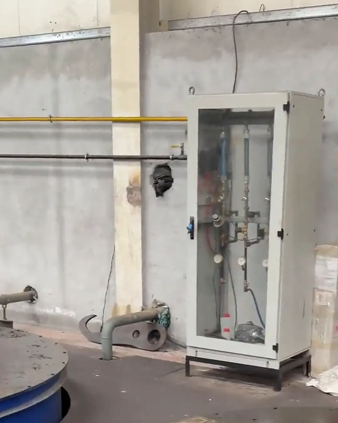 Duvar boyunca gelen boruların bağlandığı cam kapaklı beyaz dolap; içinde dikey cam tüpler, göstergeler, vanalar ve hortumlar